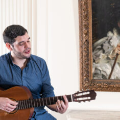 Person playing guitar in front of a painting of a portrait