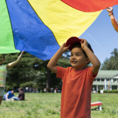 A child seen under a multicolored parachute.