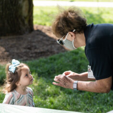 Person in a mask leaning down to talk to a child.