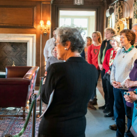 A woman stands with her back to the camera as she speaks to a group of people in a lit office.